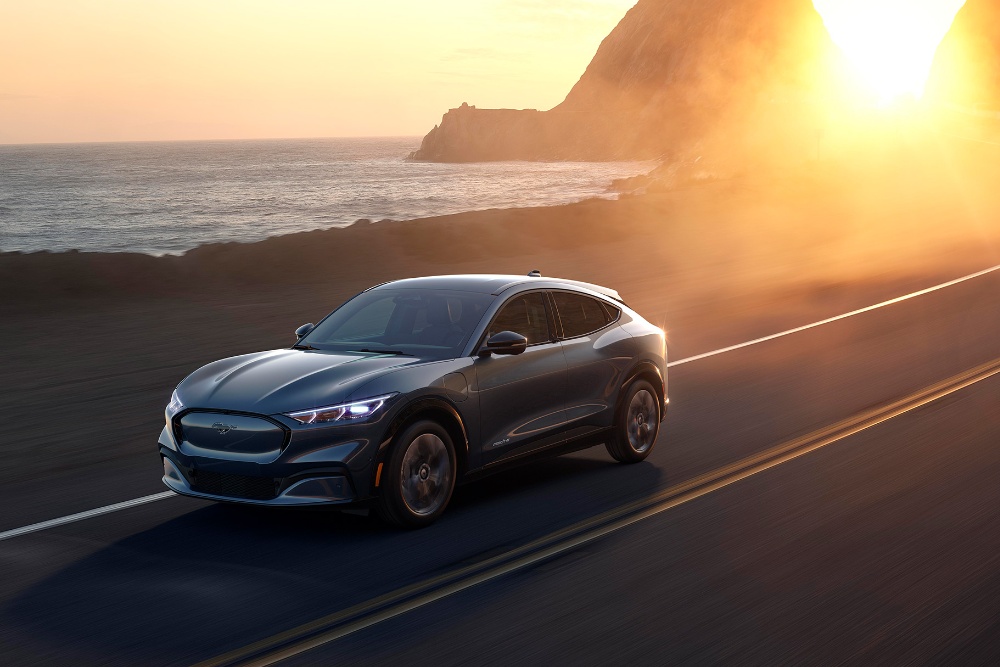 A Ford Mustang Mach-E electric car drives along a coastal road at sunset.