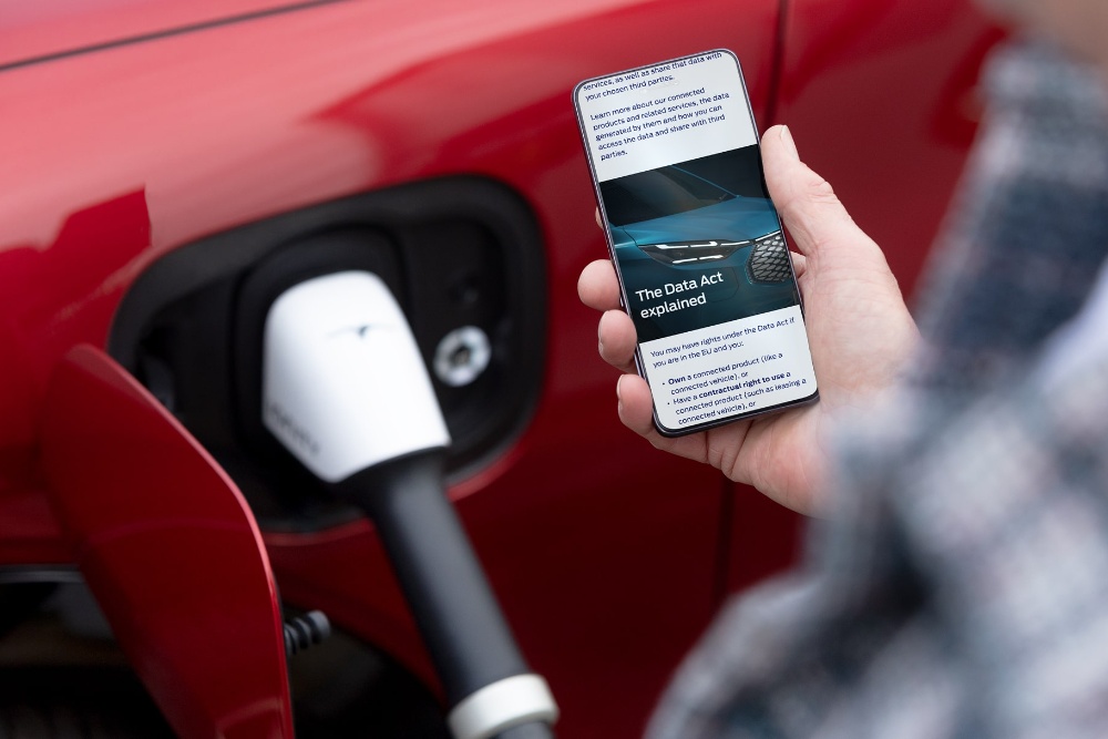 Une personne vérifiant son smartphone, un véhicule électrique rouge en charge en arrière-plan.