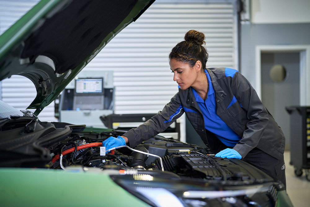 Technicienne en gants bleus travaillant sur le moteur d'une voiture Ford verte capot ouvert.