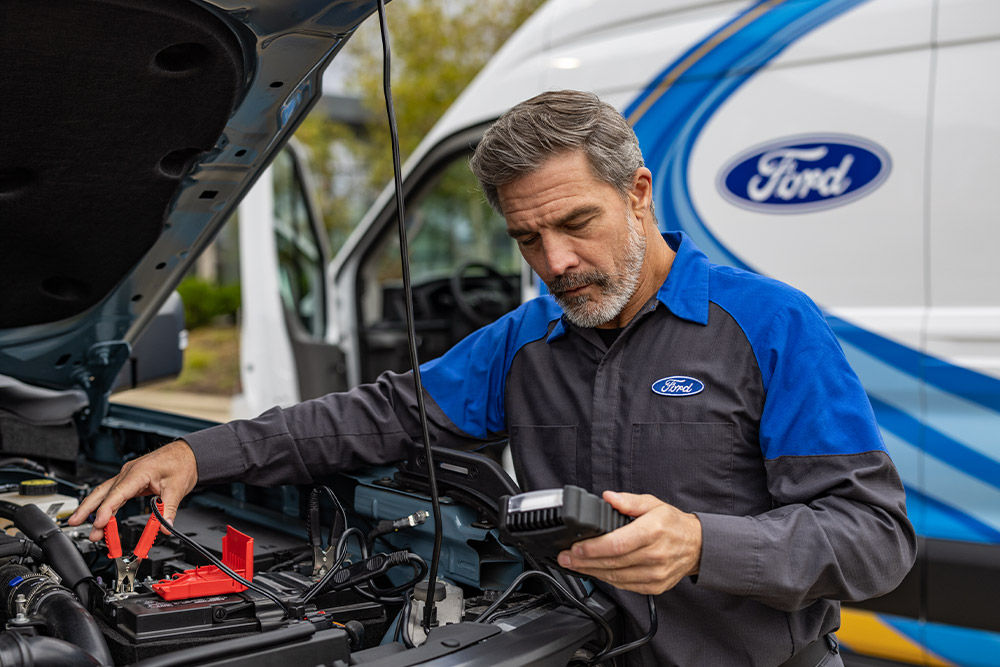 Technicien Ford en uniforme connectant des câbles de démarrage à une batterie de voiture, avec une camionnette de service Ford à proximité.