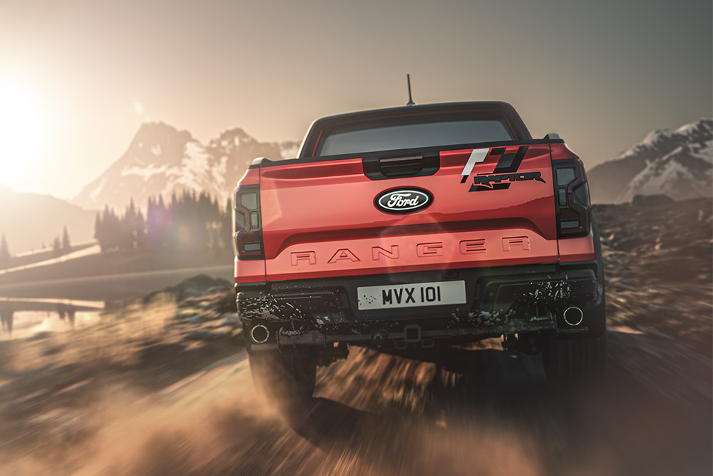 Vue arrière d’un Ford Ranger Raptor rouge roulant sur un chemin de terre avec des montagnes en arrière-plan.
