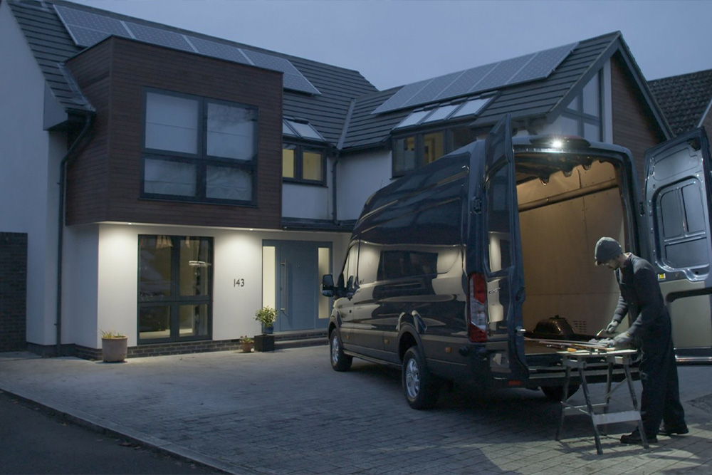Un charpentier travaille derrière un fourgon Ford Transit, portes arrière ouvertes, éclairé de nuit par le plafonnier LED.