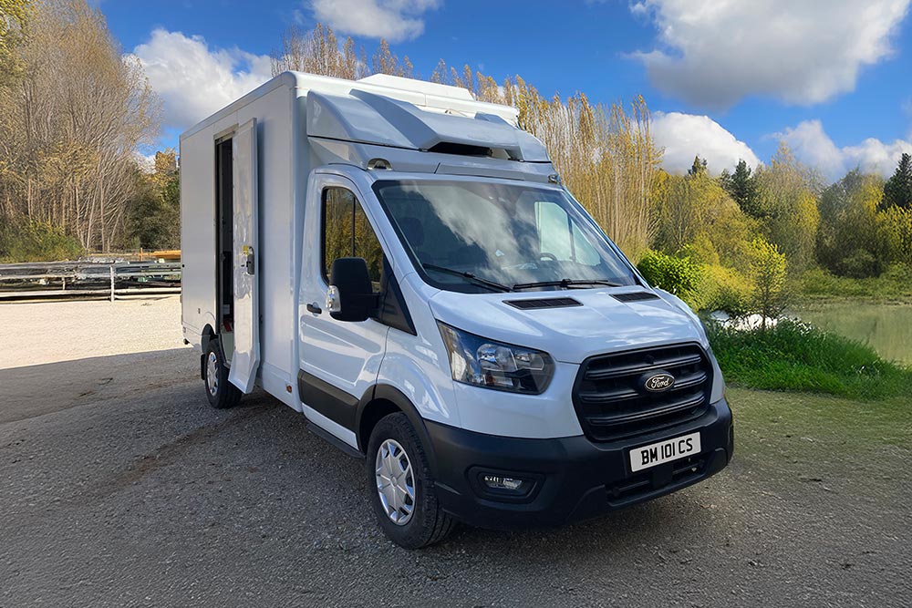 Vue 3/4 avant d'une camionnette Ford Transit blanche garée au bord d'un lac avec des arbres.