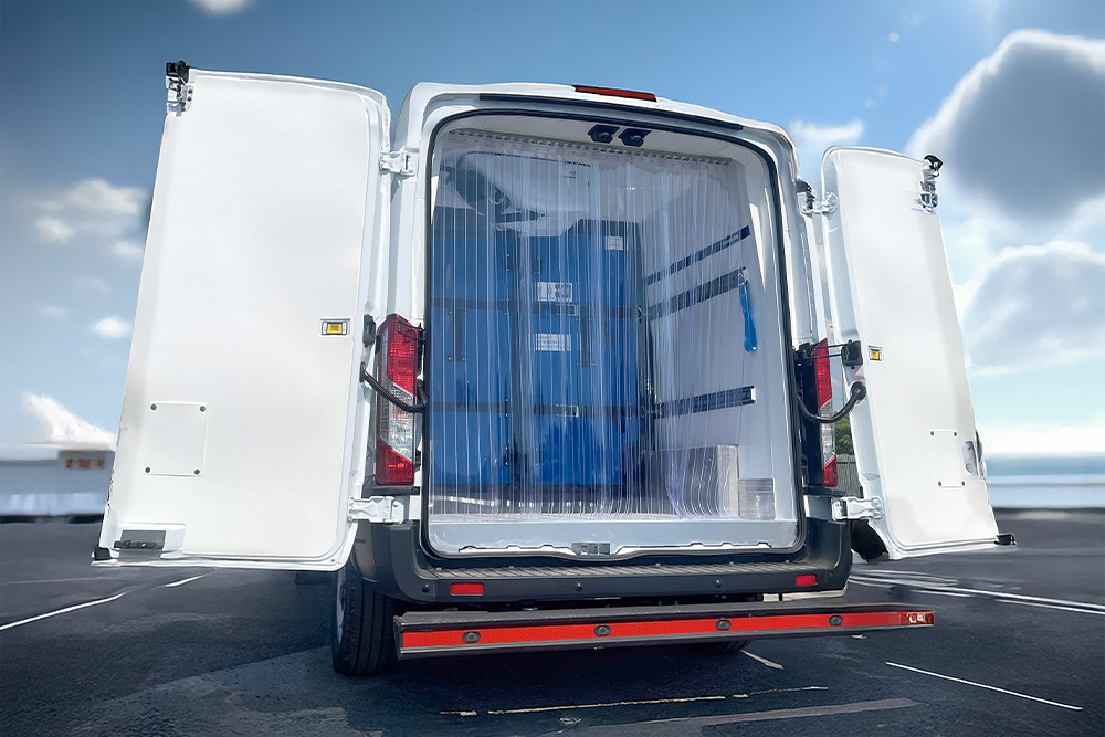 Vue arrière d'une camionnette frigorifique Ford Transit blanche avec portes ouvertes et rideaux à lanières en plastique.