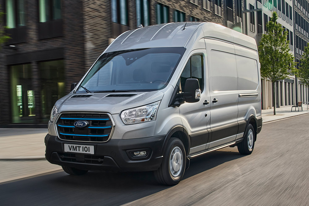 Un Ford Transit argenté circule dans un paysage urbain.