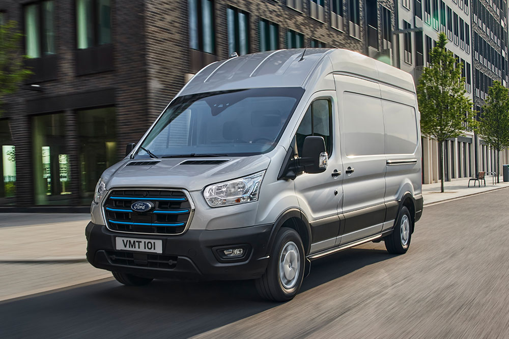 Un Ford Transit argenté circule dans un paysage urbain.