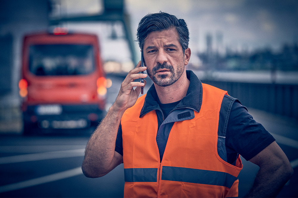 Ouvrier en gilet orange appelant l'assistance routière sur un appareil mobile.