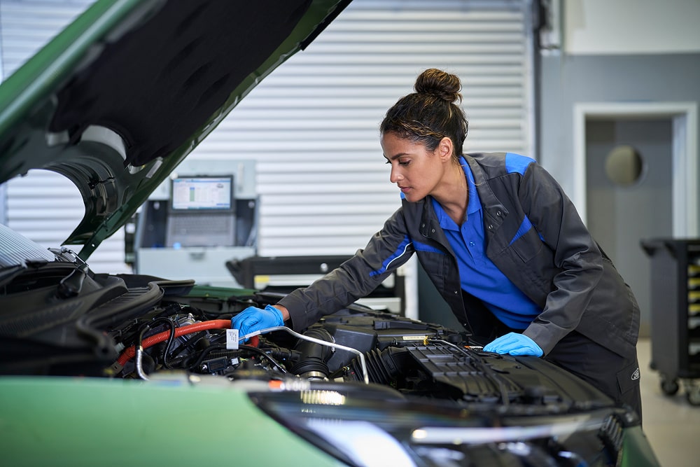 Une mécanicienne Ford inspectant un moteur de voiture avec capot ouvert dans un garage.