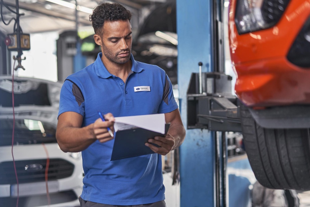 Un technicien Ford examine des documents sur un presse-papiers dans un atelier, avec une voiture sur un pont élévateur.