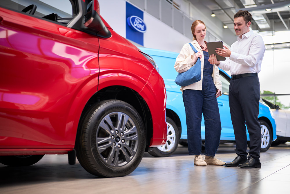 Deux personnes dans un showroom Ford regardent une tablette, avec une fourgonnette Ford rouge au premier plan et une fourgonnette Ford bleue derrière..